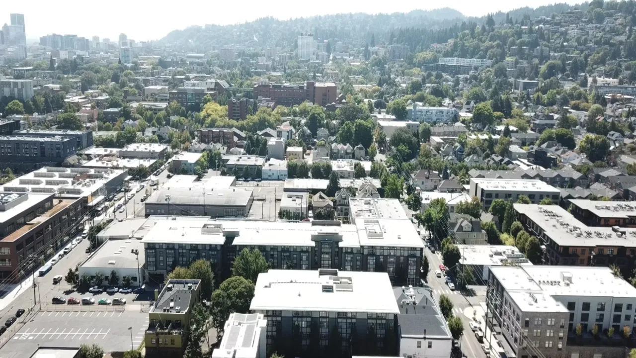 Aerial view of a cityscape with residential and commercial buildings, surrounded by lush greenery and distant hills under a clear sky.